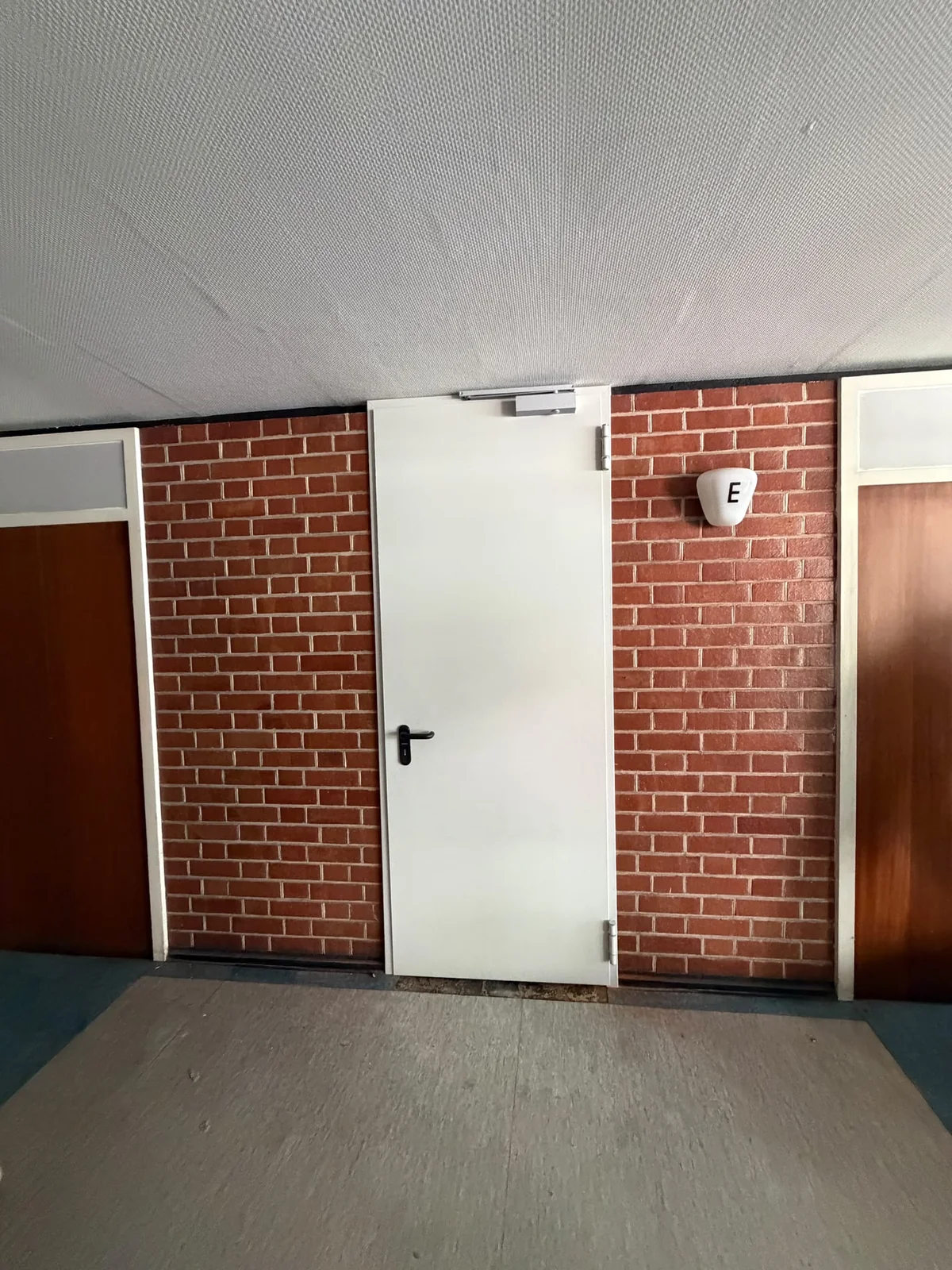 Neue weiße Tür mit Türschließer in einer Klinkerwand zwischen bestehenden braunen Türen