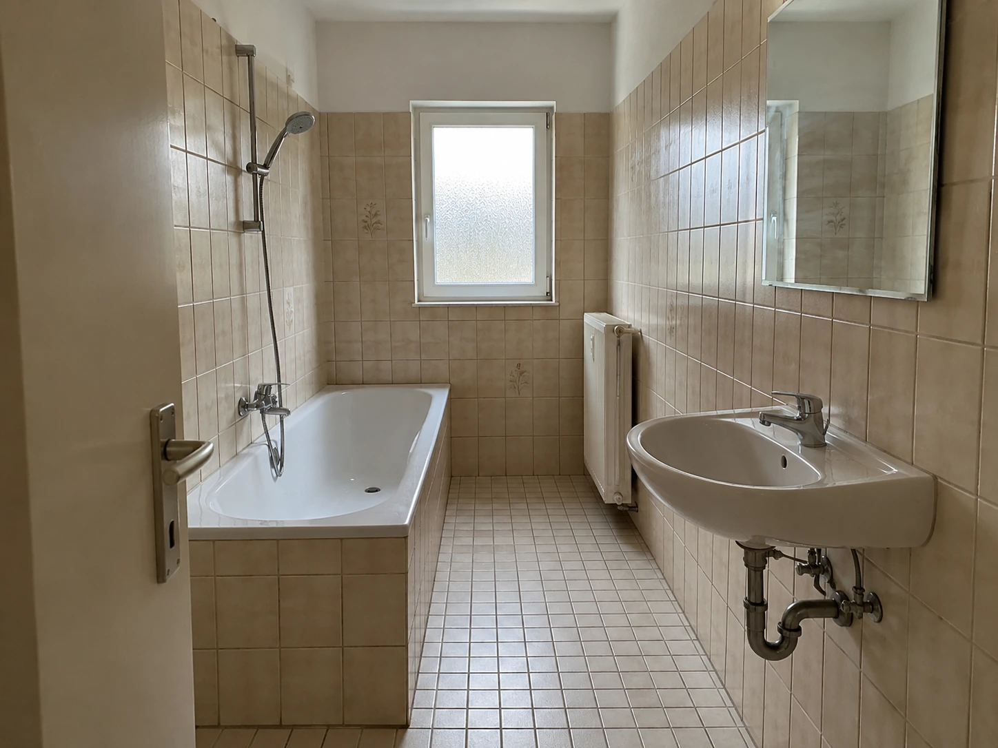 Schmales Bad mit beigen Fliesen, Badewanne links und Fenster am Raumende